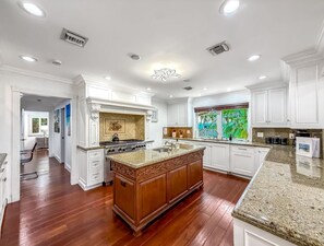 A spacious kitchen with white cabinetry, a central island, and a large window with a view of greenery. - Heated Pool & Hot Tub! Pool Table+Close To Beach! (Fort Lauderdale)