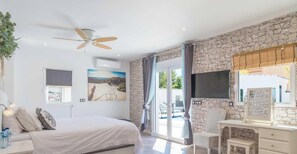 A bedroom with a bed, a desk, a TV, a ceiling fan, and a stone wall.