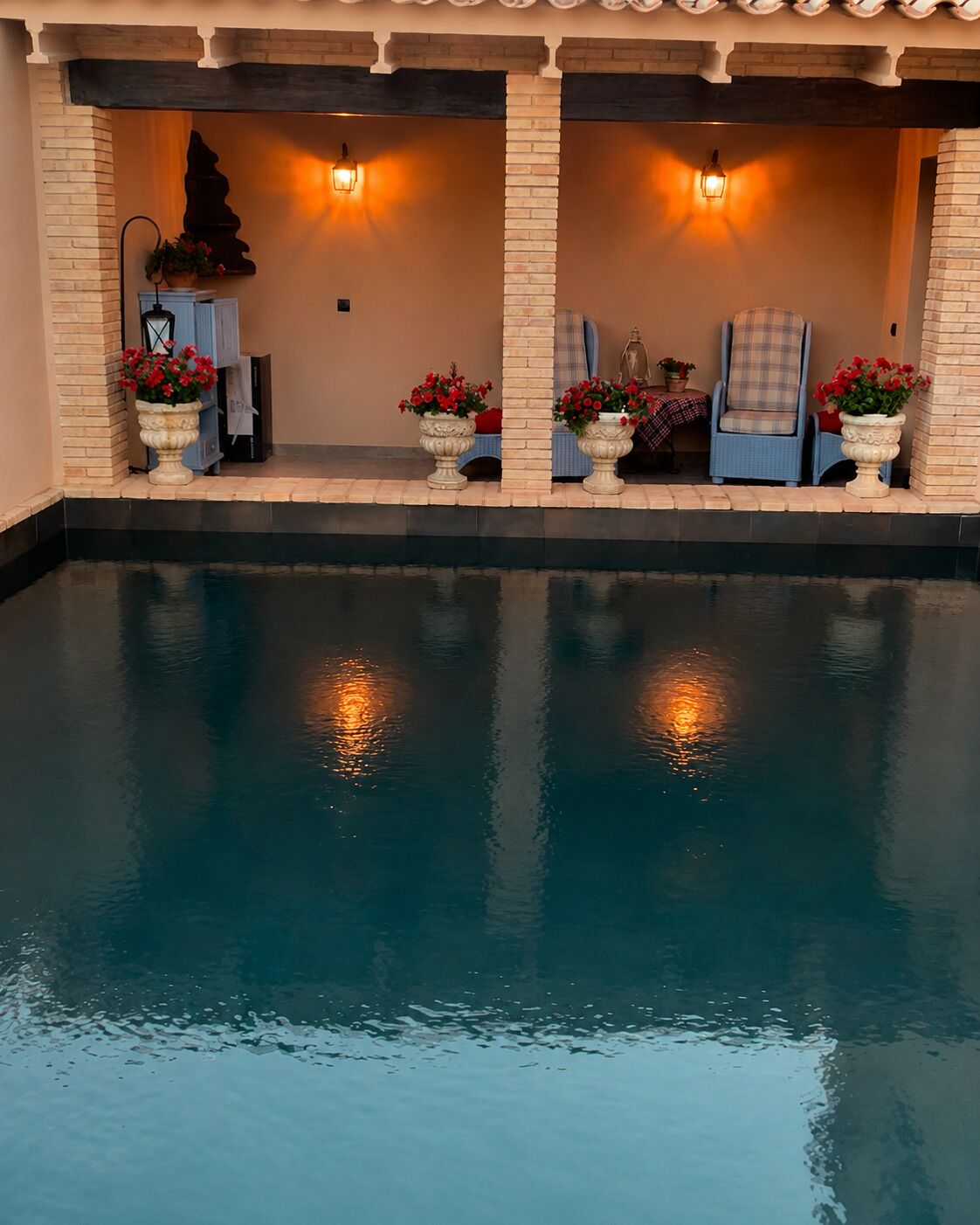 An indoor swimming pool with a seating area, two wall-mounted lights, and potted plants.