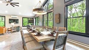 Un coin repas avec une longue table dressée pour six personnes, entourée de chaises, d’un ventilateur de plafond et de grandes fenêtres donnant sur la forêt.
