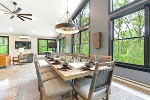 A dining area with a long table set for six, surrounded by chairs, a ceiling fan, and large windows with a forest view.