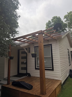Ein überdachter Patio-Bereich mit einer Holz-Pergola, einer schwarzen Tasche auf dem Deck und einem Haus mit weißem Putz und schwarzen Fensterrahmen.