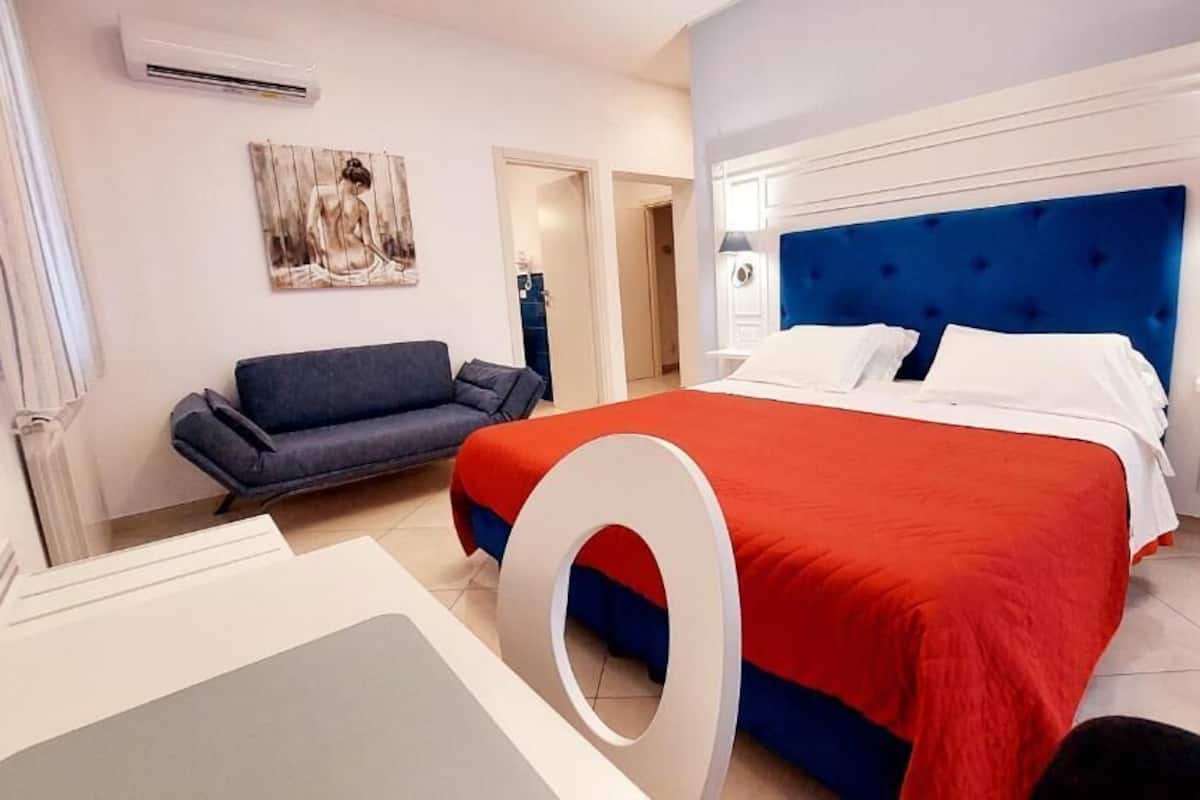 A bedroom with a bed, a sofa, a painting, and a white circular object on the floor.