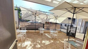 Un patio al aire libre con sillas y mesas blancas, sombrillas grandes y vistas a un edificio con balcón.