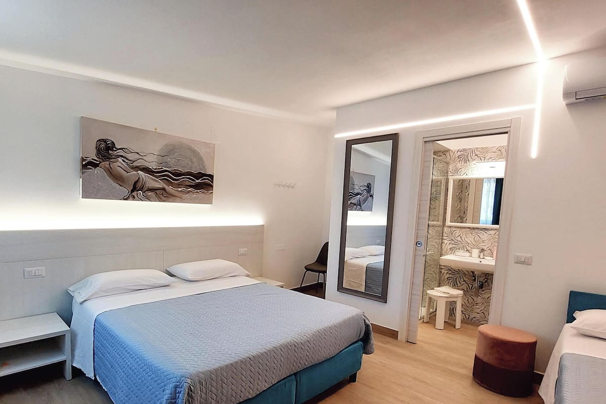 A modern bedroom with a bed, bedside tables, a mirror, a small stool, and a bathroom with a sink and mirror.