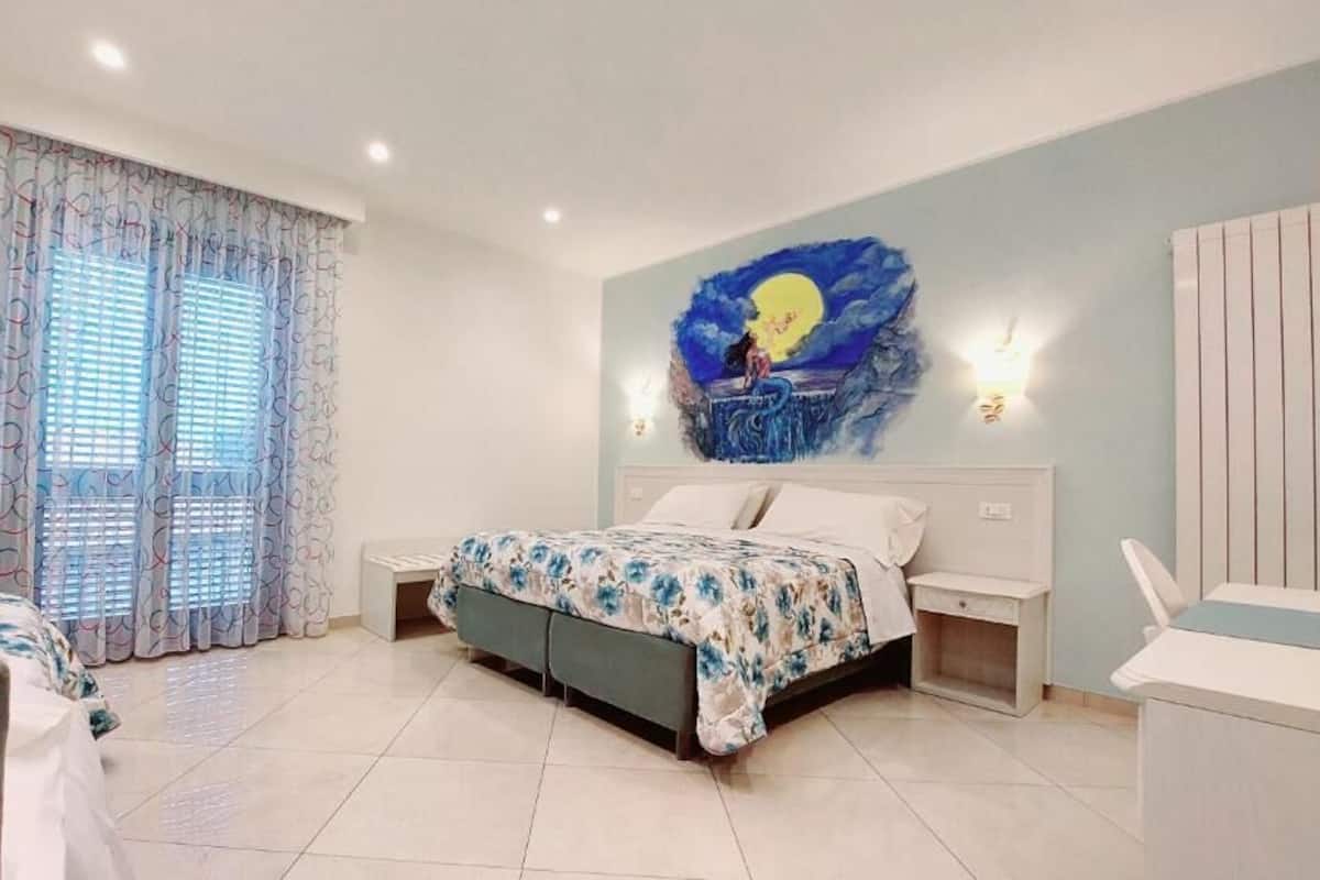 A bedroom with a bed, a nightstand, a chair, a window with curtains, and a wall painting.