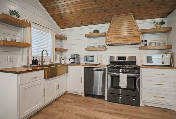 A kitchen with white cabinetry, wooden shelves, a stainless steel sink, a stove, and a microwave. - The Tenley -Boutique Cottage in Tech Terrace Walk to TTU, J&B & Goodline (Lubbock)