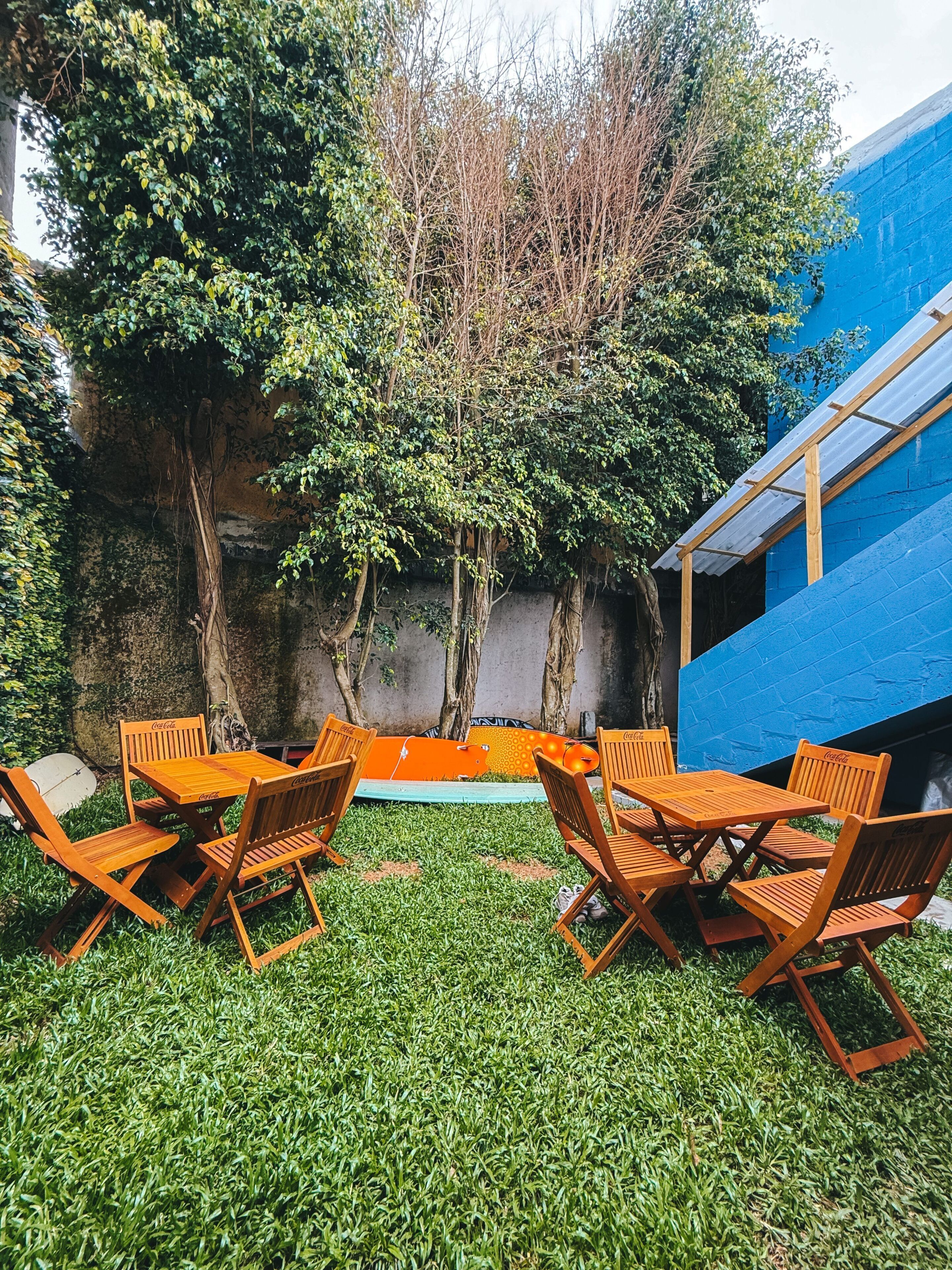 A patio with wooden chairs and a table, surrounded by lush greenery and a blue wall.