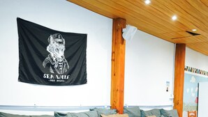 A modern living room with a wooden ceiling, a sofa, a coffee table, and a wall-mounted flag. - Sea Wolf Surf Hostel - Campeche (Florianópolis)