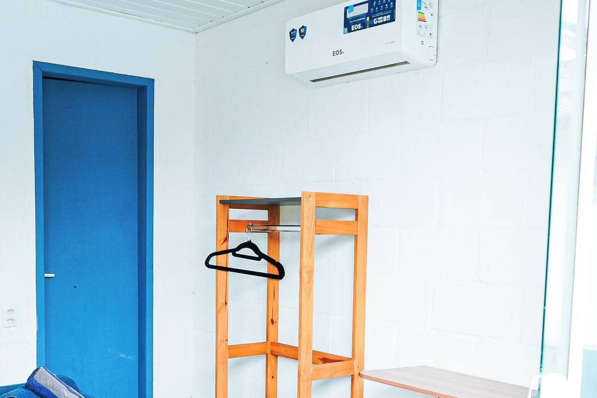 Una habitación con cama, puerta azul, una silla de madera, estante y unidad de aire acondicionado.