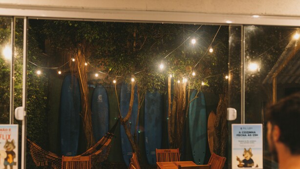 A cozy outdoor seating area with string lights, wooden furniture, and a view of trees through a glass wall. - Sea Wolf Surf Hostel - Campeche (Florianópolis)