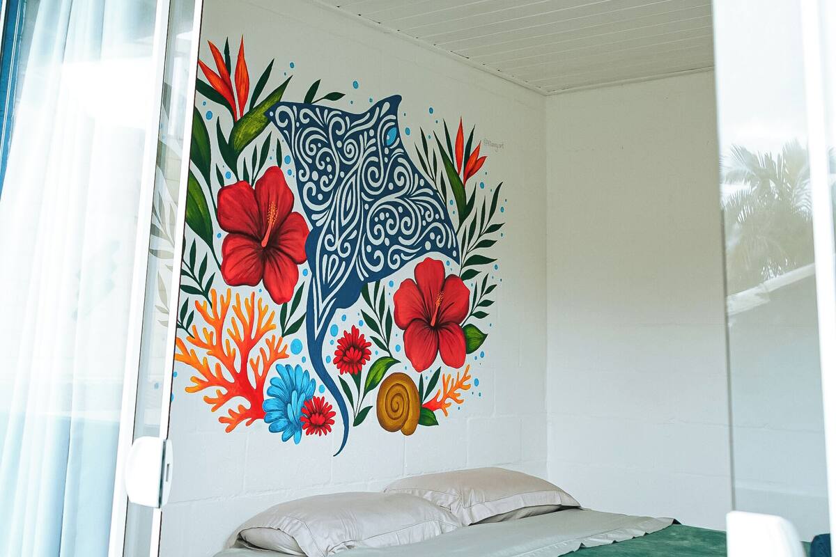 Una habitación con una cama, un ventanal con vistas y una pared decorada con un mural floral de colores.