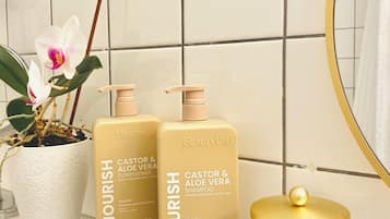 Ein Badezimmerschrank mit zwei Flaschen Nourish Kastanien- und Aloe-Vera-Spülung und Shampoo, einer Topf-Orchidee und einem Glasbehälter mit goldfarbenem Deckel.