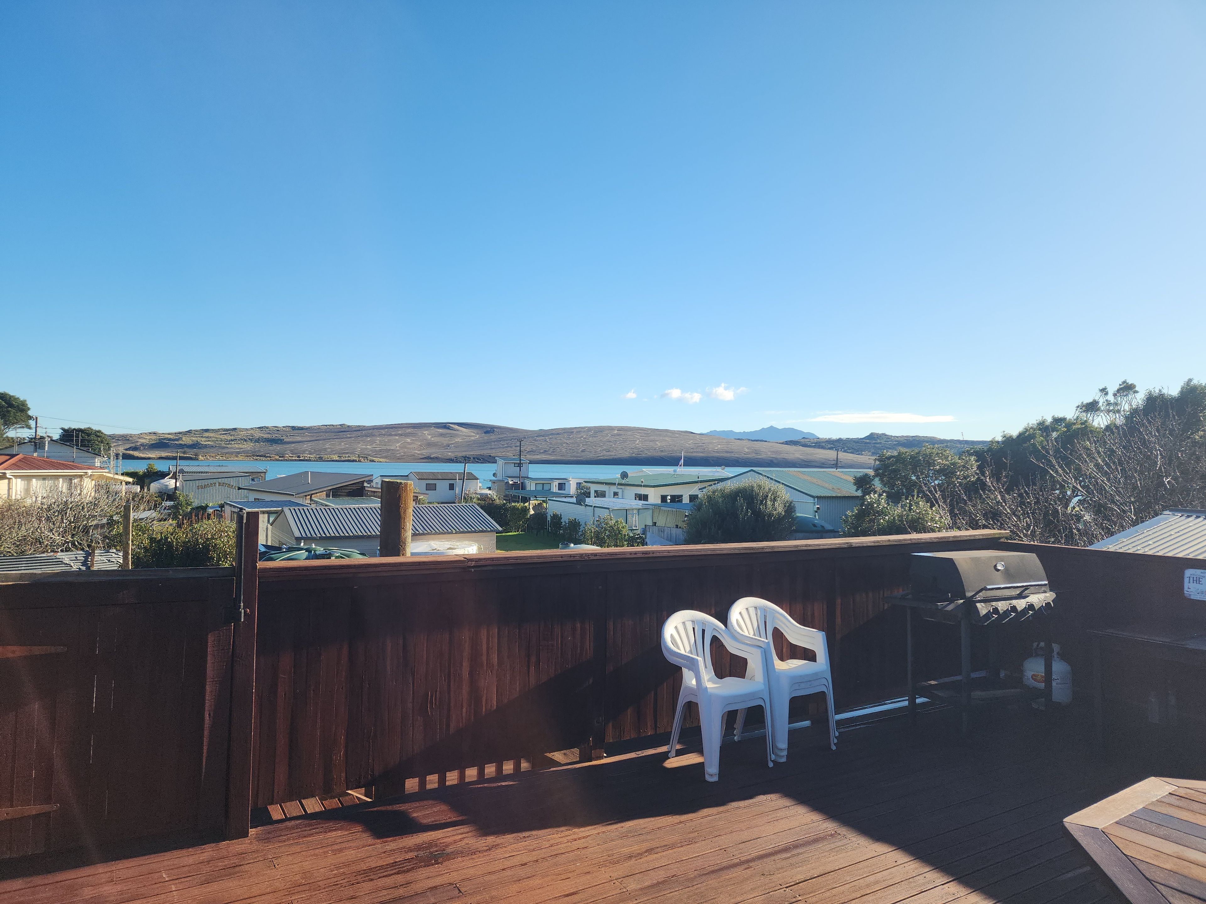 A wooden deck with a barbecue grill and two white plastic chairs, overlooking a residential area with houses and a body of water.