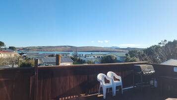 A wooden deck with a barbecue grill and two white plastic chairs, overlooking a residential area with houses and a body of water.