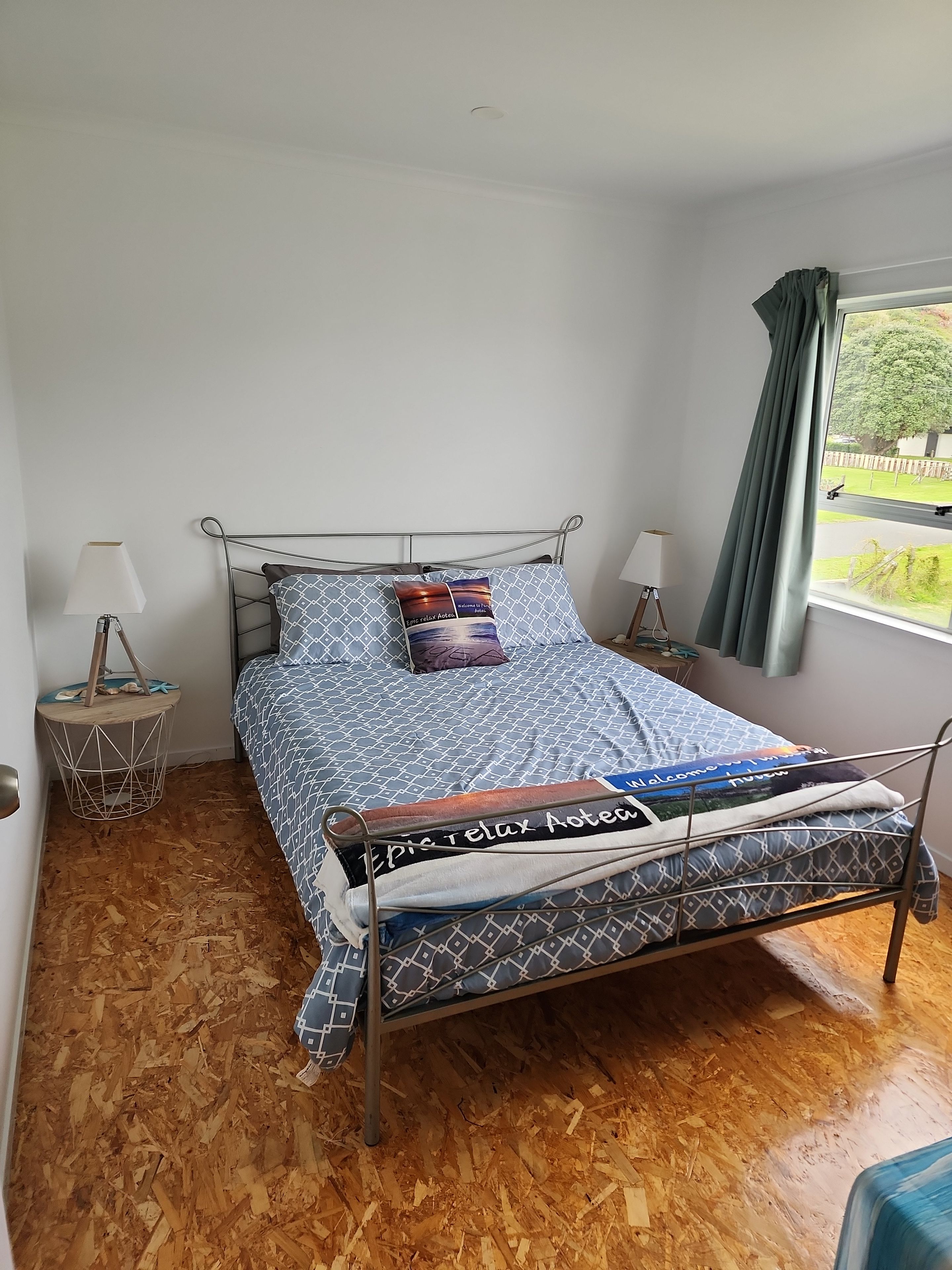 Un lit simple avec un cadre en métal, une couvre-lit à motifs et des oreillers. Il y a deux tables de chevet avec des lampes, et une fenêtre avec des rideaux.