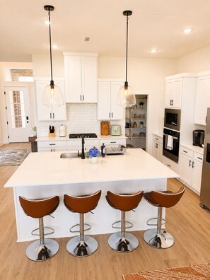 A modern kitchen with white cabinets, a large island with bar stools, stainless steel appliances, and pendant lighting. - Noah's Landing: New 4BR Sleeps 9, Near UofA, AMP, Hospital & Trails. Pets Ok (Springdale)