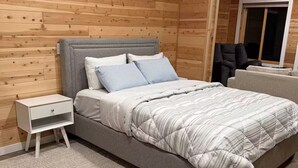 A bedroom with a wooden wall, a grey bed, a white nightstand, and a patterned rug. - Cedar Studio w Full Kitchen + Living @ Whitetail Estates (Babbitt)