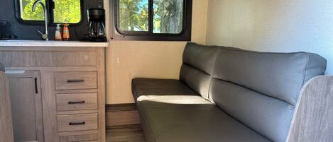 A compact RV interior with a kitchenette, a cushioned seating area, and a window providing a view of trees.