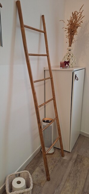 Eine Holzleiter lehnt an einem weißen Schrank, ein Korbwesen enthält eine Toilettenpapierrolle, und auf dem Schrank steht eine Vase mit Trockenpflanzen.