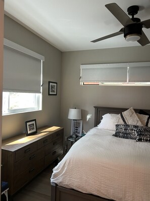 Ein Schlafzimmer mit Bett, Nachttisch, Kommode, Deckenventilator und Fenster mit Jalousien.