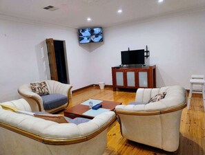 Ein Wohnzimmer mit zwei weißen Ledersofas, einem Holztisch, einem an der Wand montierten Fernseher und einem Schrank mit einem Flachbildfernseher.