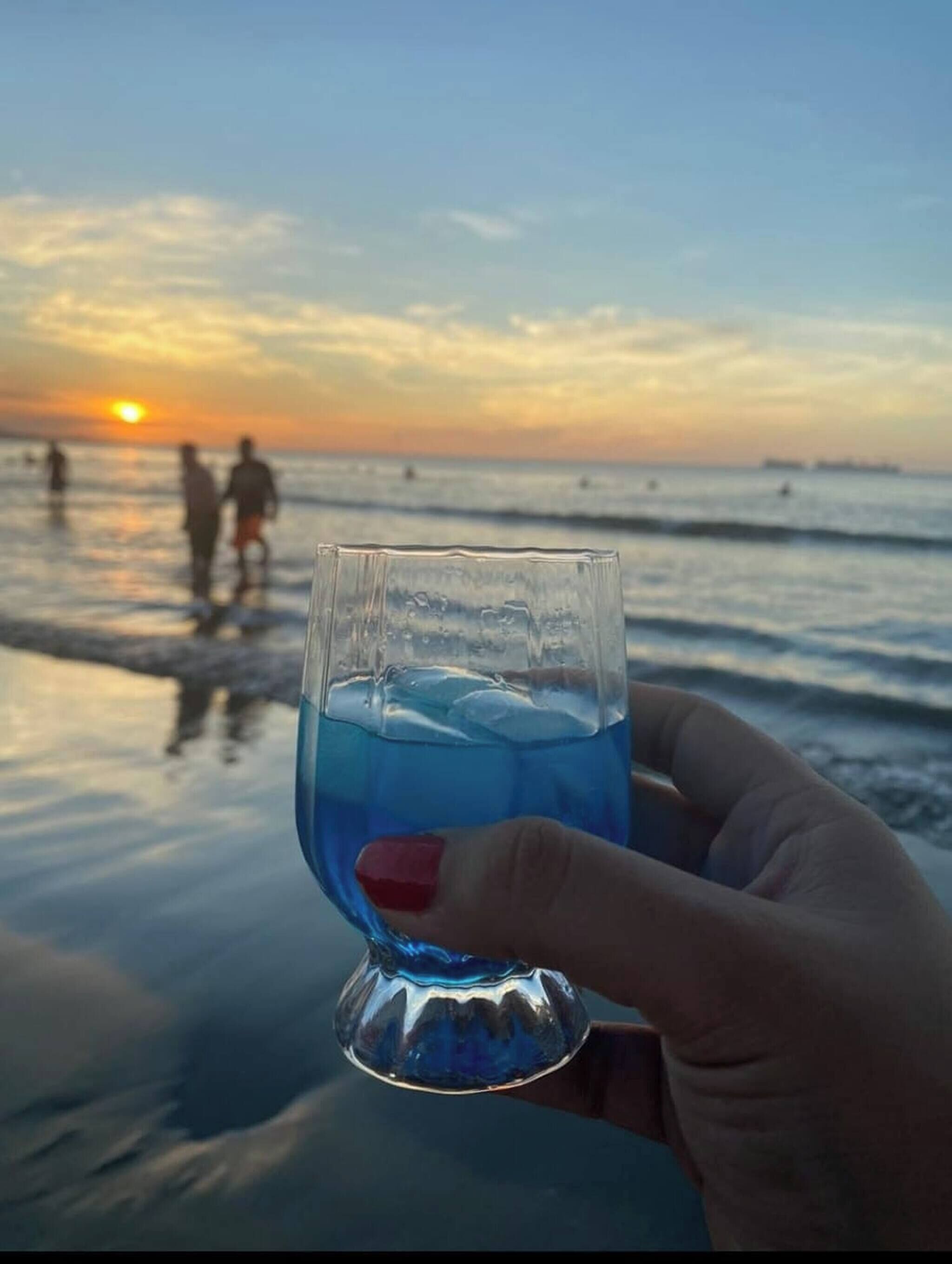 Una mano che tiene un bicchiere con liquido blu, sullo sfondo di una spiaggia al tramonto.