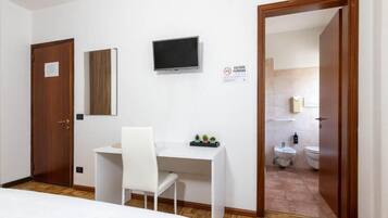 A hotel room with a bed, a desk with a chair, a television, a door, and a bathroom visible through an open door.