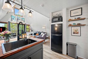 Una cocina con un refrigerador negro, gabinetes de madera y un lavamanos negro. Hay cuadros en la pared y una repisa de madera con tazas.