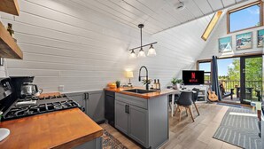 A modern kitchen with a gas stove, a coffee maker, and a sink. There is a dining area with a table and chairs, and a TV mounted on the wall.