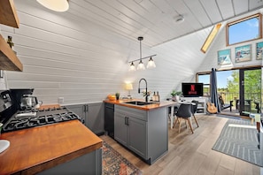 A modern kitchen with a gas stove, a coffee maker, and a sink. There is a dining area with a table and chairs, and a TV mounted on the wall.