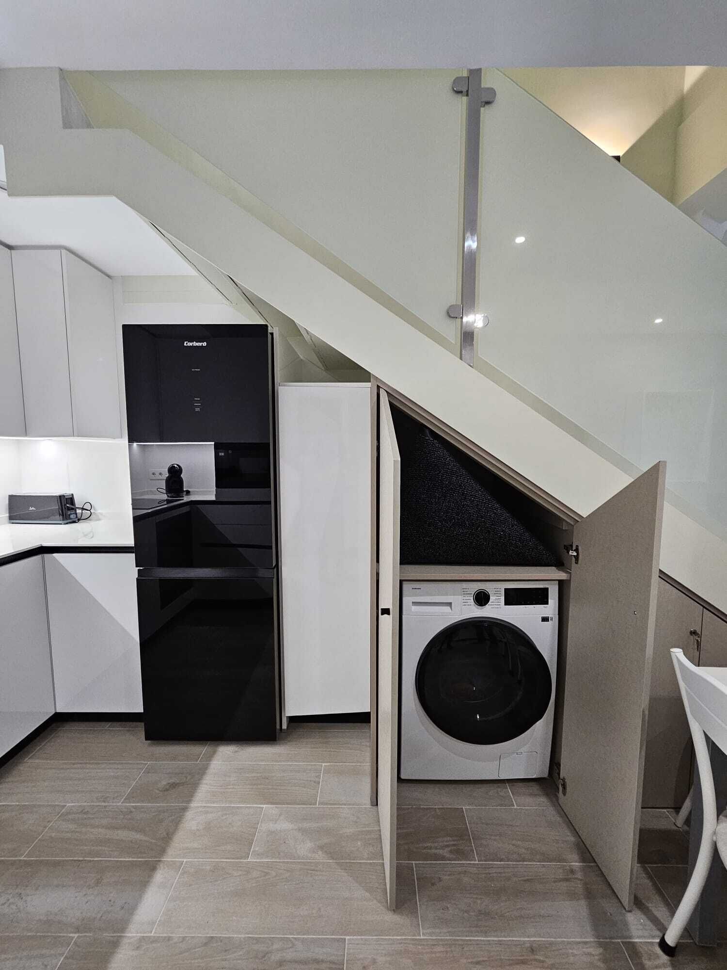 Une cuisine compacte équipée d’un lave-linge et d’un sèche-linge intégrés, d’un réfrigérateur et d’un escalier menant à un niveau supérieur.