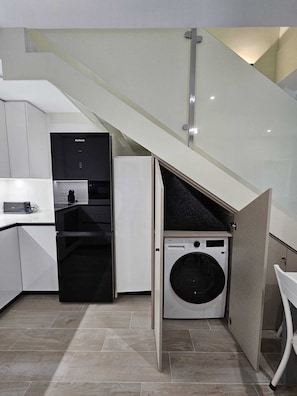 A compact kitchen with a built-in washer and dryer, a refrigerator, and a staircase leading to an upper level. - La Herencia B. Fully equipped duplex. (Ponferrada)