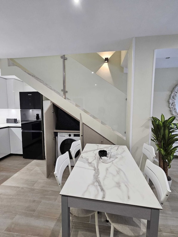 A modern kitchen with a built-in washing machine, a dining area with a marble table, and a staircase with a glass railing.