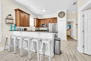 A modern kitchen with wooden cabinets, a stainless steel refrigerator, and a built-in microwave. There are white bar stools and a clock on the wall. - Destin Beach Vibes, sleeps 13, 2min walk to private beach, heated community pool (Destin)