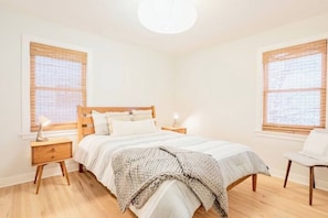 Ein Schlafzimmer mit einem Holzbett, zwei Nachttischen, einem Sessel und Fenstern mit Jalousien.