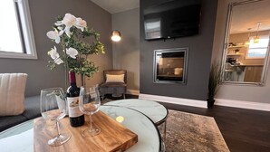 A modern living room with a glass table, a wine bottle, and glasses. There is a TV mounted on the wall and a fireplace in the background. - Chic Stay in Wortley – Walk to Downtown & Parks (London)