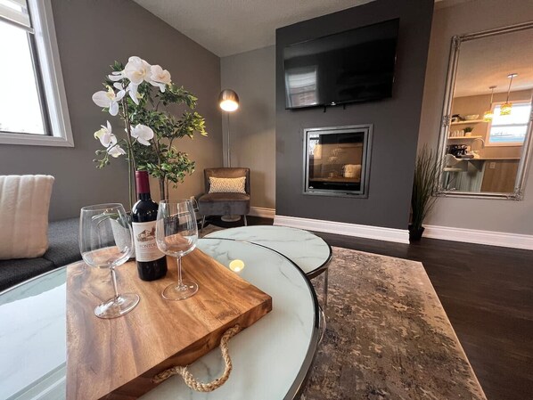 A modern living room with a glass table, a wine bottle, and glasses. There is a TV mounted on the wall and a fireplace in the background.