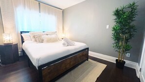 A bedroom with a bed, a nightstand, a potted plant, and a window with curtains.
