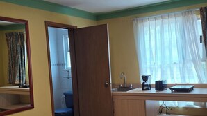 Une pièce comprenant un lit, une salle de bain visible à travers une porte ouverte, un lavabo avec un miroir et une fenêtre avec des rideaux.