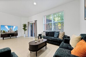 Ein modernes Wohnzimmer mit einem großen Flachbildfernseher, einem dunklen Holztisch, einem dunkelgrauen Sofa mit vielen Kissen, einem großen Fenster mit Jalousien und einer Topfpflanze.