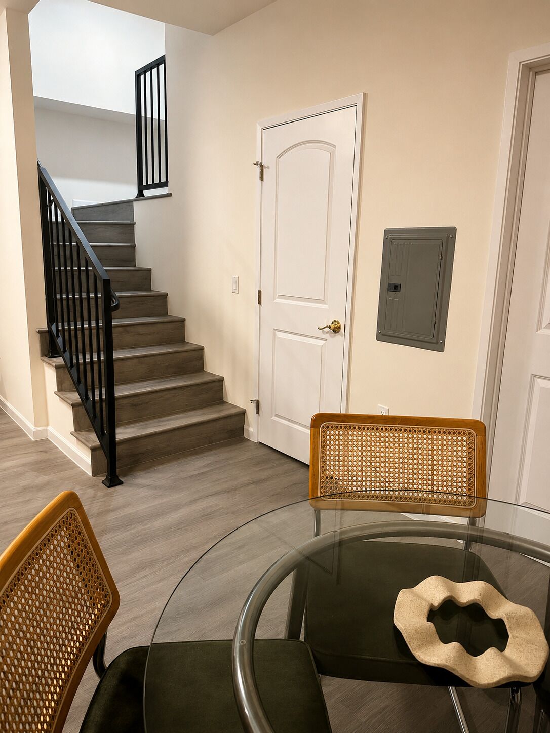 Un escalier avec des rampes noires menant à une porte blanche, une table ronde en verre avec une chaise, et une chaise en osier.