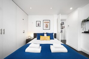 A modern hotel room with a large bed, a wardrobe, a nightstand, and a wall-mounted shelf with decorative items. - Chic Arlington Home | Walk to Angel & King’s Cross (Greater London)