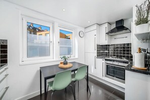 A modern kitchen with white cabinets, a black stove, and a dining area with a table and chairs. - Chic Arlington Home | Walk to Angel & King’s Cross (Greater London)