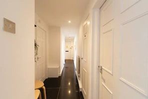 A narrow hallway with white doors, a small table, and a plant on the left wall. - Chic Arlington Home | Walk to Angel & King’s Cross (Greater London)