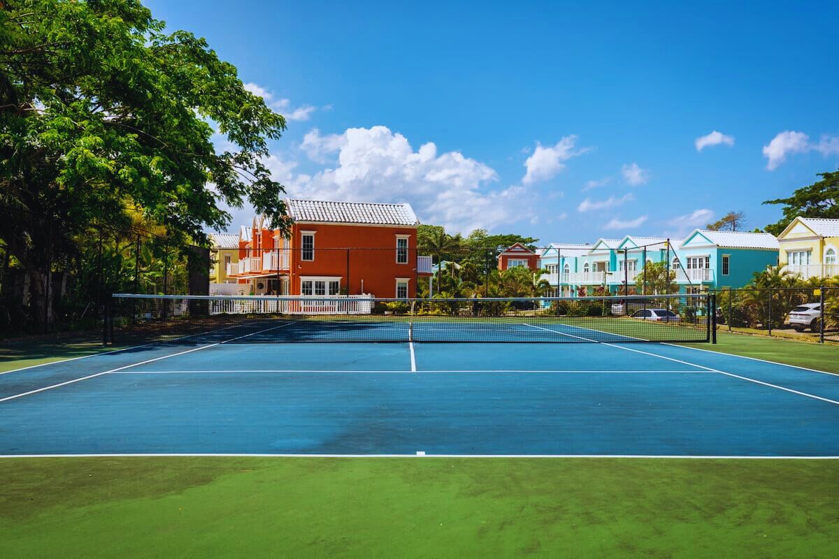 En tennisbane med net, omgivet af farverige huse, træer og en klar himmel.