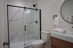 A bathroom with a glass shower enclosure, a toilet, a sink with a mirror, and a towel rack. - Maison Atelier | Centrally Located | Designer 1bd (Los Angeles)