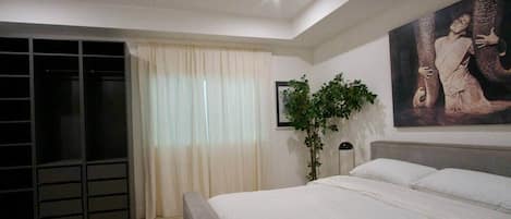 A bedroom with a bed, a large window with curtains, a plant, and a painting on the wall.