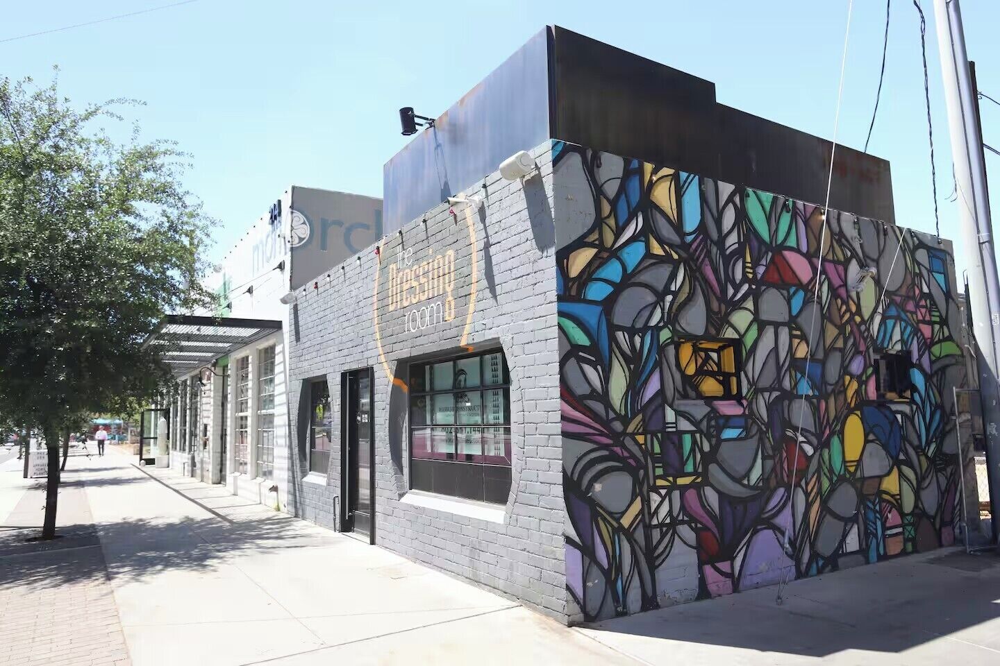 Un edificio con un mural abstracto y colorido en una de sus fachadas, una tienda con un letrero que dice 'The Design Room', y una acera con árboles y un cielo despejado.