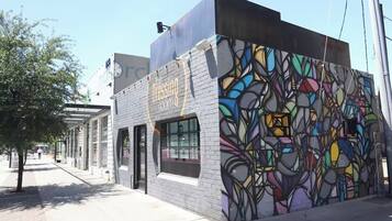 Un edificio con un mural abstracto y colorido en una de sus fachadas, una tienda con un letrero que dice 'The Design Room', y una acera con árboles y un cielo despejado.